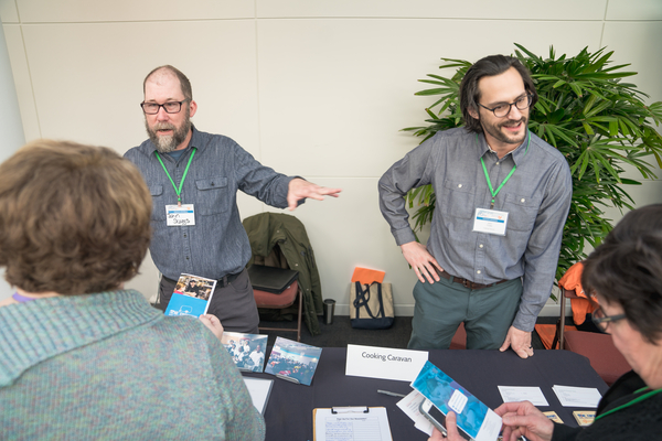 Two men wearing name badges standin behind a table talking with a group of people.