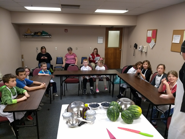 Six children sit at tables. Between them is another table covered with white paper. On top are watermelons, bowls and other utensils