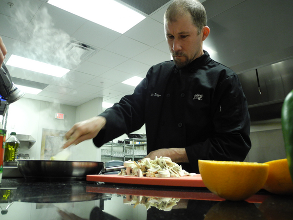 A chef stirs food in a saute pan, next to a pile of chopped mushrooms.